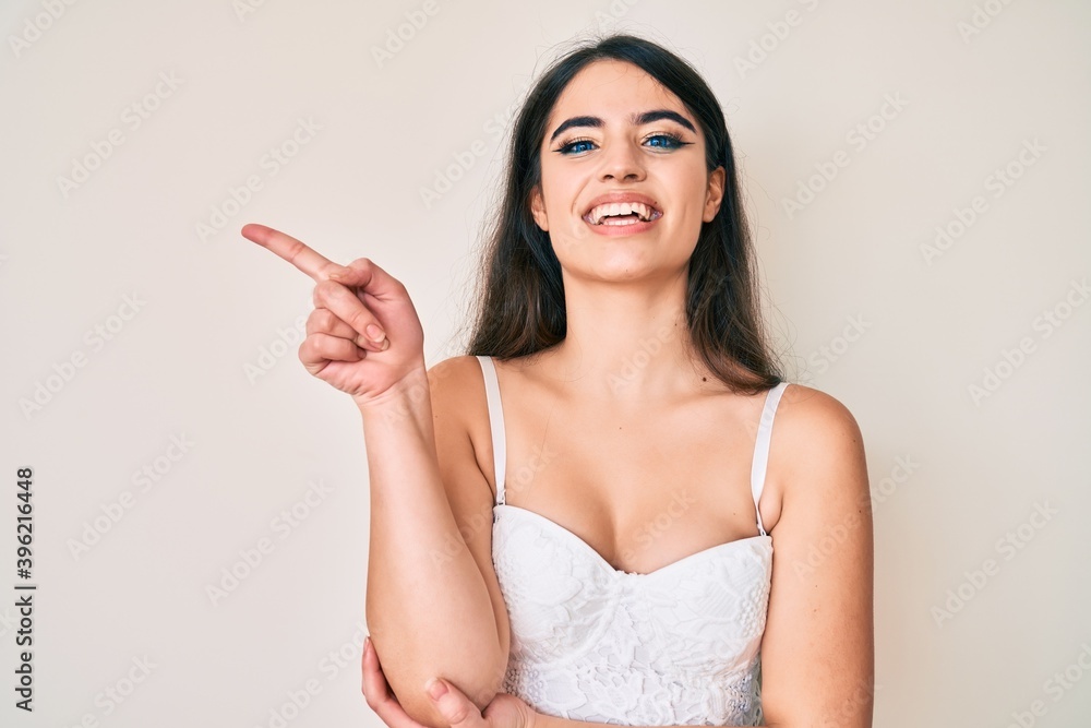 Brunette teenager girl posing elegant with a big smile on face, pointing with hand and finger to the side looking at the camera. Brunette teenager girl posing elegant with a big smile on face, pointing with hand and finger to the side looking at the camera.