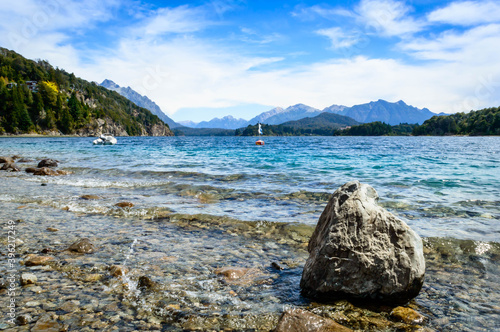 Beautiful landscape seen from the ground behind a large rock. Crystal clear water, mountains, pine trees and rocks. Beach in Bariloche, Argentina on a very sunny day. photo