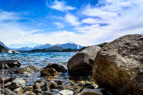 Beautiful landscape seen from the ground behind a large rock. Crystal clear water, mountains, pine trees and rocks. Beach in Bariloche, Argentina on a very sunny day. © Rariel