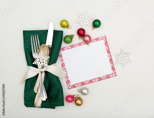 Christmas Place Setting with Silverware in Green Napkin on Off White Table Cloth Background with menu card and colorful ornaments.