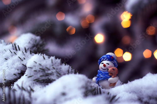 Happy smiling snowman in knitted clothes, with a little dog under the arm is standing in snowy spruce forest landscape, with warm unfocused lights on cold background,merry christmas and happy new year
