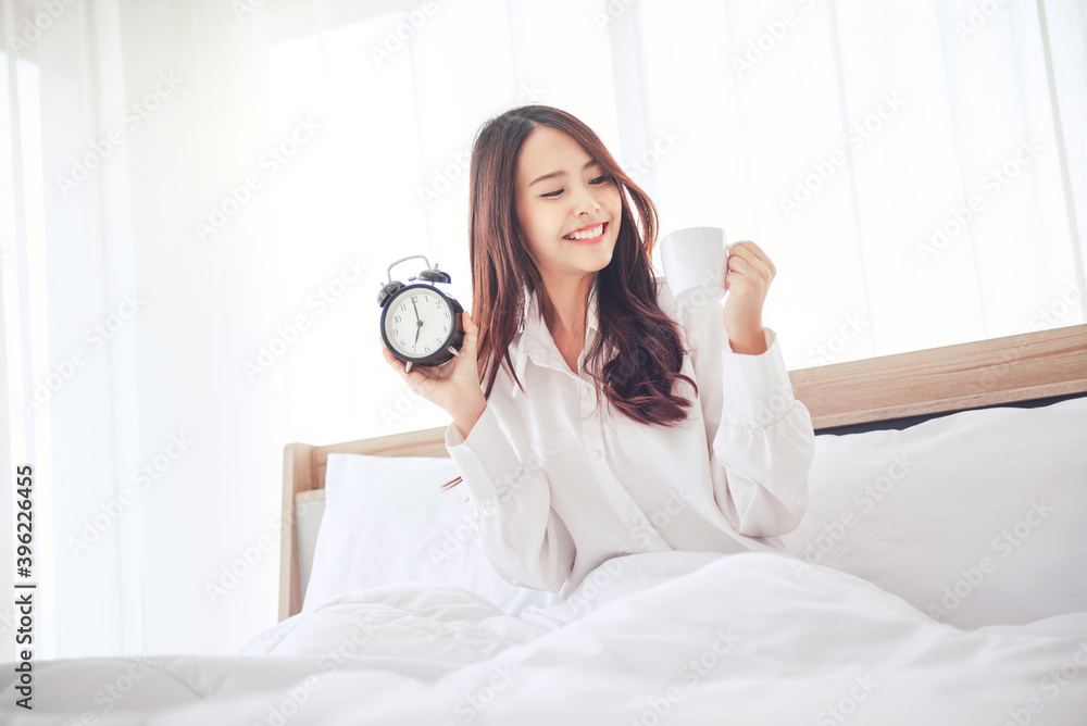Happy asian woman holding alarm clock and a coffee cup on the bed in morning. Happy asian woman holding alarm clock and a coffee cup on the bed in morning.