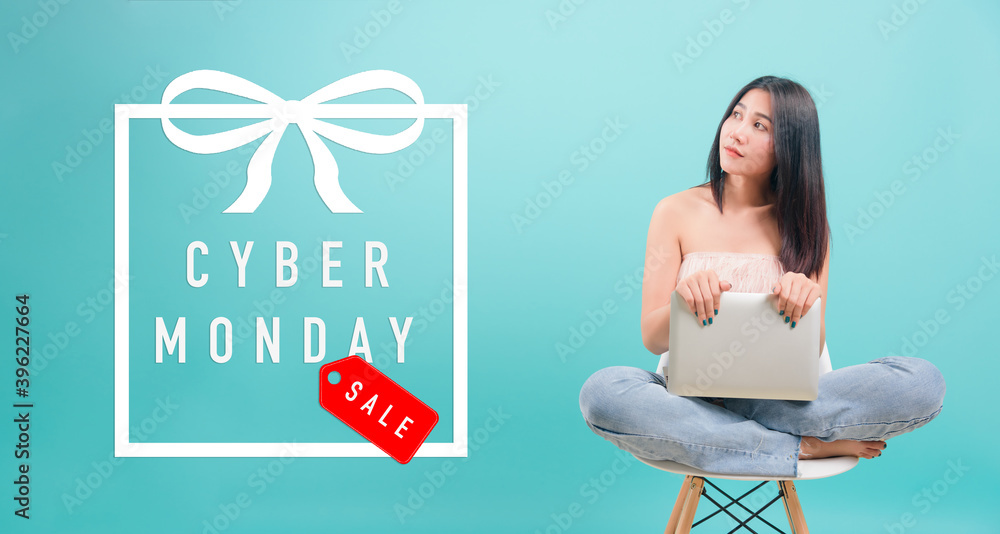 Asian happy portrait beautiful young woman sitting on chair smile her  holding laptop computer and looking to side with Cyber Monday text in gift box on blue background
