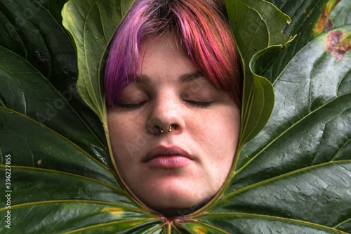 Portrait of a young woman with pink hair with eyes closed, sleepy and dreaming face expression surrounded by a big green natural leaf