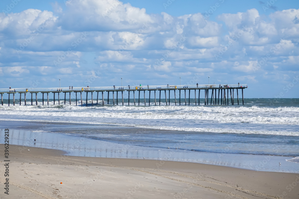 Long pier extending from the beach out into a rough ocean. The sky is blue with many white puffy clouds Long pier extending from the beach out into a rough ocean. The sky is blue with many white puffy clouds