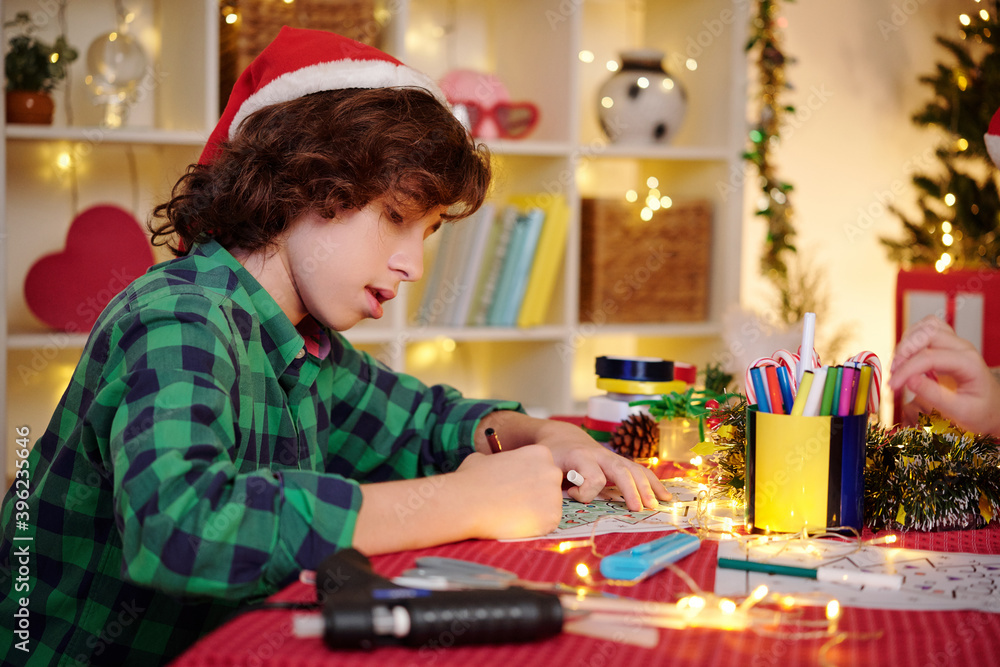 Talented creative Hispanic boy in Santa Claus hat drawing Christmas related picture at table