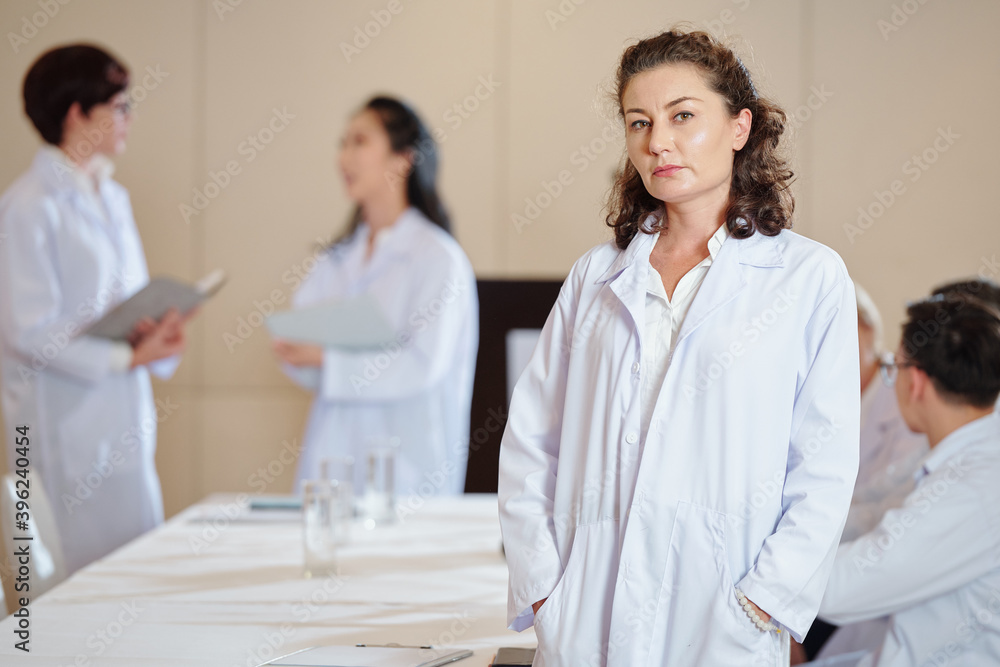 Serious mature female pharmacologist in labcoat standing at table in boardroom and looking at camera Serious mature female pharmacologist in labcoat standing at table in boardroom and looking at camera