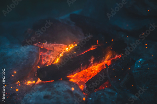 Vivid smoldered firewoods burned in fire close-up. Atmospheric background with orange flame of campfire. Unimaginable full frame image of bonfire. Glowing embers in air. Warm logs, bright sparks bokeh