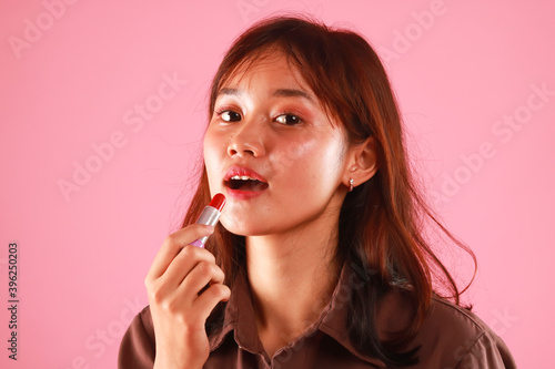 Portrait of young girl painting her lips with bright lipstick on pink background.