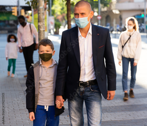 Man in disposable face mask walking with his preteen son along city street. Concept of Covid 19 virus spread prevention and human safety