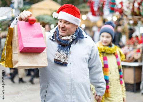 Portrait of smiling man in Santa hat with bags while shopping on Christmas street fair with his teen daughter