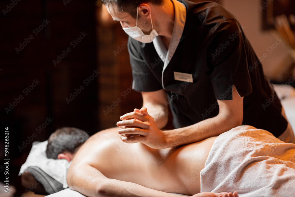 Professional masseur in facial mask doing a deep massage to a male client at Spa salon. Business during the epidemic concept