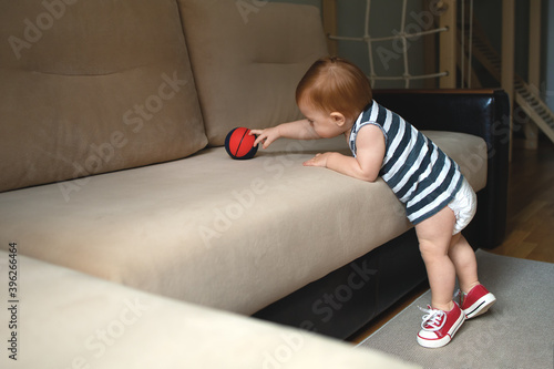 a purposeful kid reaches for the ball standing on his toes near the sofa indoors, a sporty kid is dressed in a striped sleeveless T-shirt, red sneakers and a dry white diaper