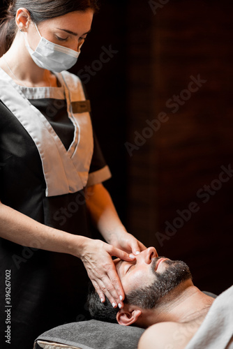 Professional female masseur in medical mask doing facial massage to a male client at Spa salon. Business during the epidemic concept