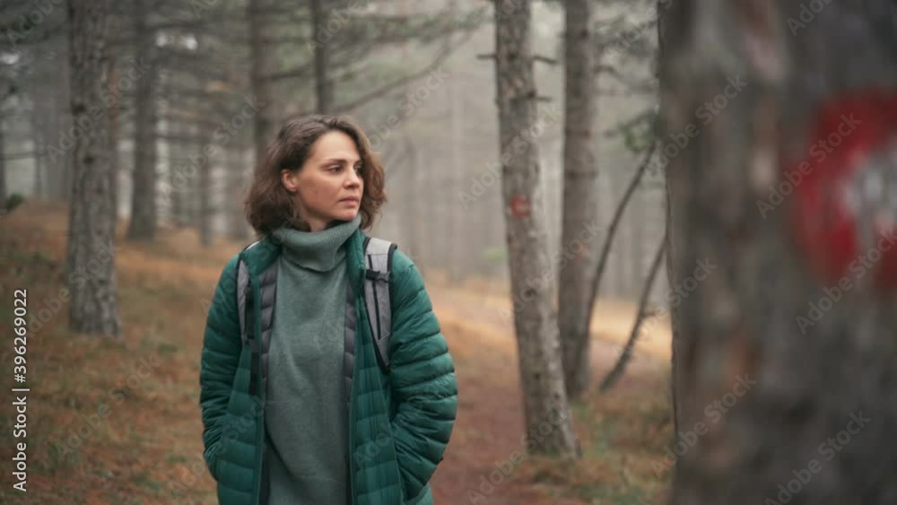A young woman with a backpack walks along a trail in the autumn misty forest and passes by a tree with a red route mark. A young woman with a backpack walks along a trail in the autumn misty forest and passes by a tree with a red route mark.