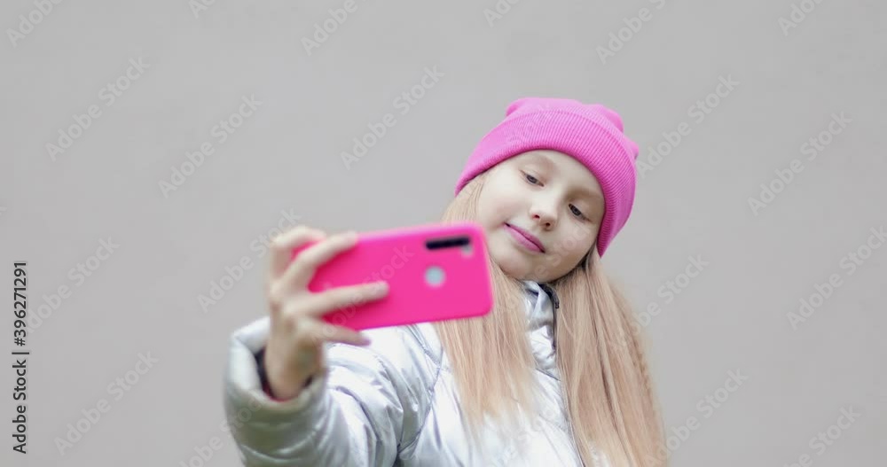 A fashionable schoolgirl in a pink hat makes a selfie outdoors. Girl holding a mobile phone in her hands. A fashionable schoolgirl in a pink hat makes a selfie outdoors. Girl holding a mobile phone in her hands.