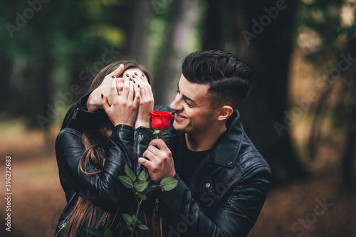 Handsome brunette man covers woman's eyes by hand and gives her red rose, in autumn park.
