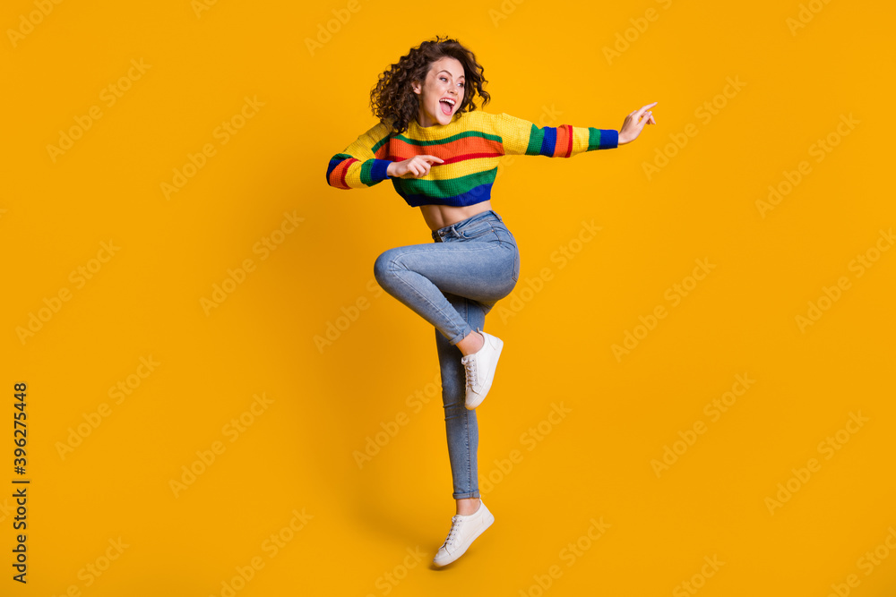 Full length photo of jumping lady wearing casual colorful sweater dancing pointing looking empty space standing one leg isolated yellow color background