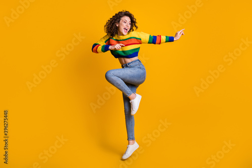 Full length photo of jumping lady wearing casual colorful sweater dancing pointing looking empty space standing one leg isolated yellow color background