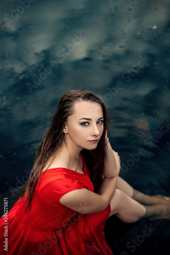 Vertical portrait of young pretty wet woman mermaid sitting on pier rock in the water river or lake touching head, dressed in red dress, looking at the camera, copy space and nature blur background.