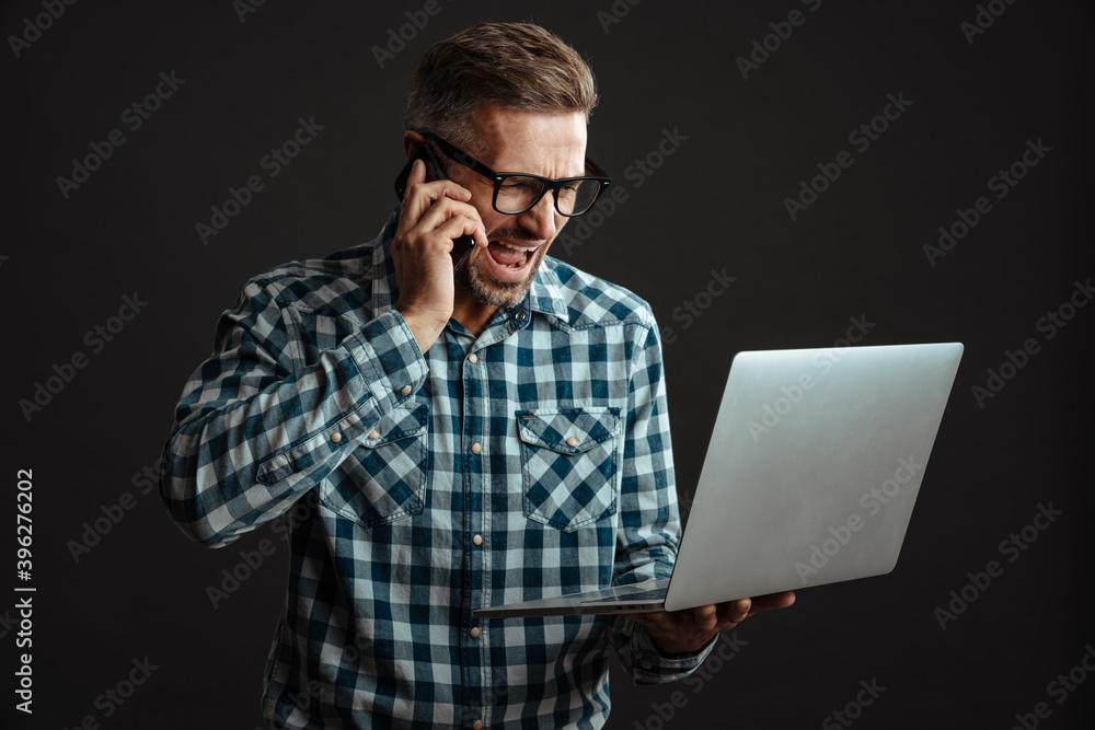 Irritated man in eyeglasses holding laptop and talking on cellphone Irritated man in eyeglasses holding laptop and talking on cellphone