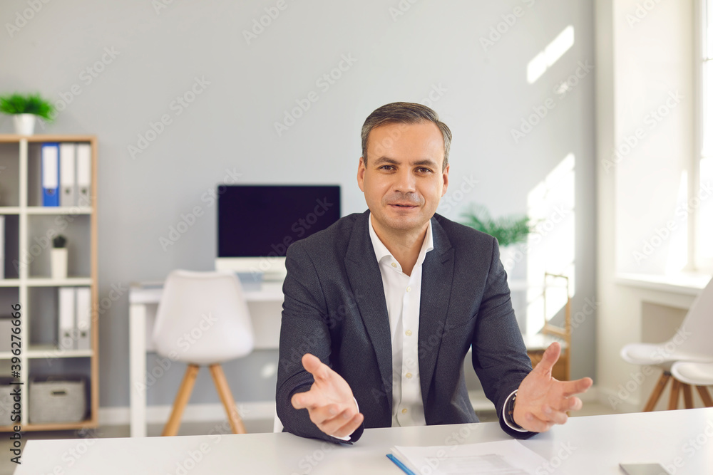 Portrait of happy businessman sitting at desk in office, looking at camera and talking