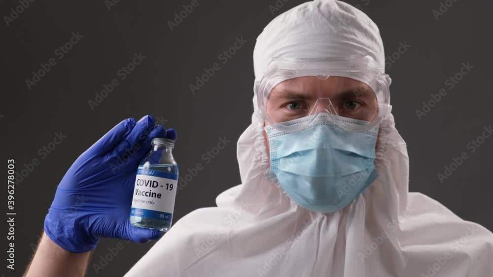 Scientist in a protective suit, mask, glasses and gloves shows a developed vaccine against Covid. Concept of global pandemic positive end. Scientist in a protective suit, mask, glasses and gloves shows a developed vaccine against Covid. Concept of global pandemic positive end.