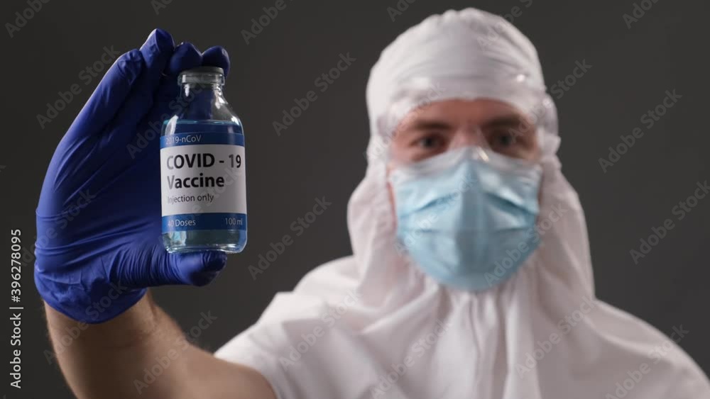 Scientist in a protective suit, mask, glasses and gloves shows a developed vaccine against Covid. Concept of global pandemic positive end. Scientist in a protective suit, mask, glasses and gloves shows a developed vaccine against Covid. Concept of global pandemic positive end.