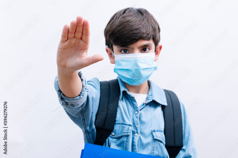 Handsome child boy student with backpack and mask holding books on isolated white background with open hand making stop sign Handsome child boy student with backpack and mask holding books on isolated white background with open hand making stop sign