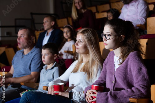 Friendly family of four sitting with popcorn and drinks in cinema, watching movie with interest. Family leisure and entertainment concept © JackF