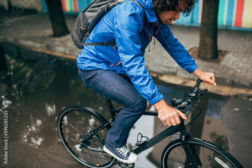 Horizontal image of a handsome man cycling on his bike down the street next to the house. Male courier with curly hair delivers parcel cycling with a bicycle in the city on a rainy day.