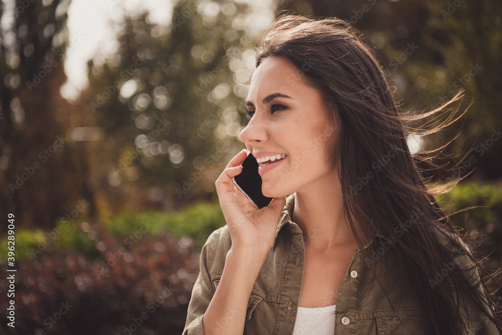 Photo of lovely dreamy young woman look side hold telephone speak toothy smile wear brown shirt park street outside Photo of lovely dreamy young woman look side hold telephone speak toothy smile wear brown shirt park street outside