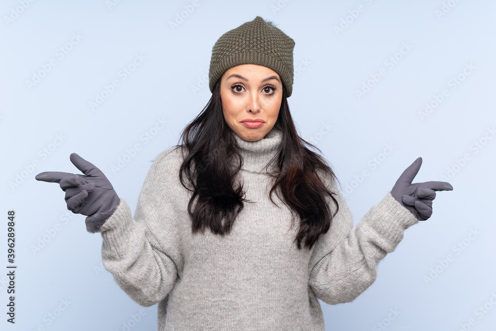 Young Colombian girl with winter hat over isolated blue background pointing to the laterals having doubts Young Colombian girl with winter hat over isolated blue background pointing to the laterals having doubts