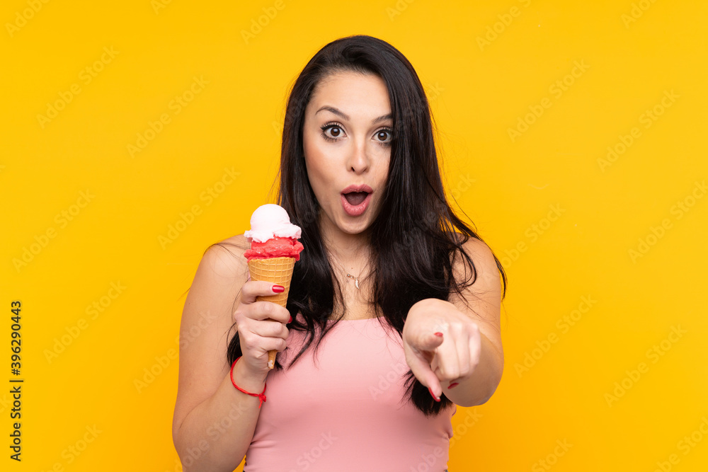 Young Colombian girl holding an cornet ice cream over isolated yellow background surprised and pointing front Young Colombian girl holding an cornet ice cream over isolated yellow background surprised and pointing front