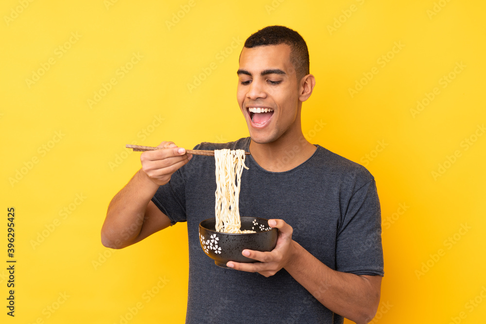 Young African American man over isolated yellow background holding a bowl of noodles with chopsticks and eating it Young African American man over isolated yellow background holding a bowl of noodles with chopsticks and eating it