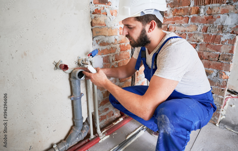 Young plumber, wearing blue uniform and white helmet working with sealant fix of sewer pipe in kitchen or in bathroom in unfinished apartment. Concept of plumbing works and home renovation Young plumber, wearing blue uniform and white helmet working with sealant fix of sewer pipe in kitchen or in bathroom in unfinished apartment. Concept of plumbing works and home renovation