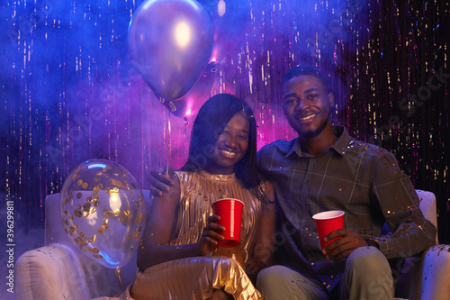 Portrait of young African-American couple smiling at camera while sitting on couch against sparkling background and enjoying party, copy space