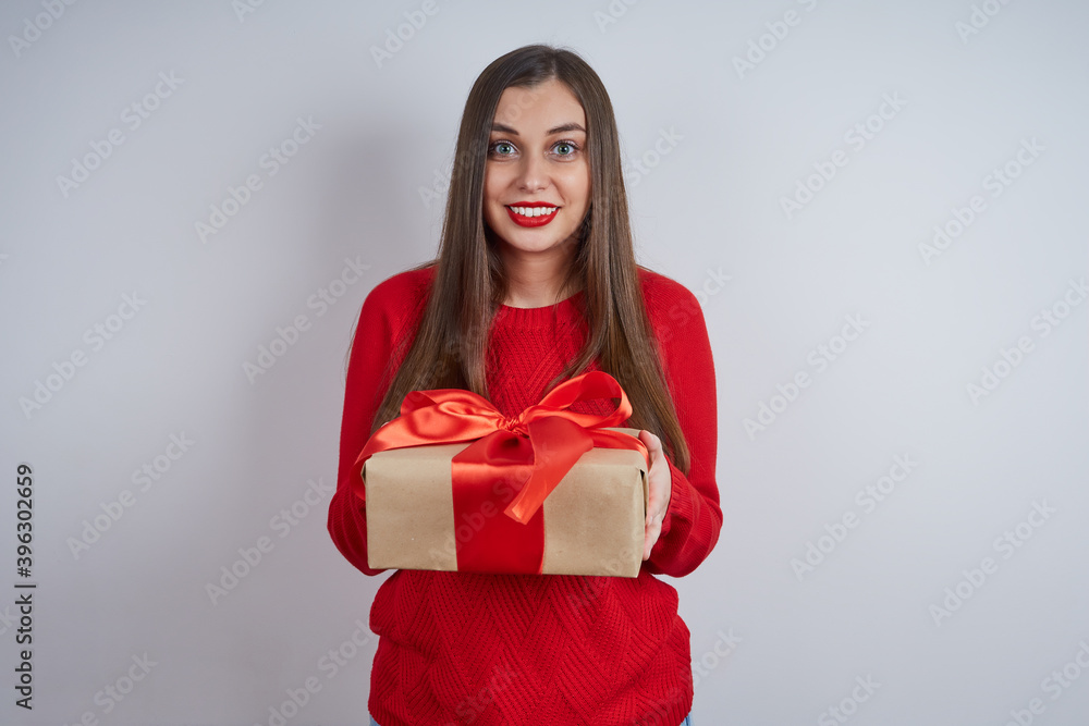 Beautiful surprised woman in a red sweater, holding a gift box, looking at the camera on a gray background. The concept of holiday, Christmas, New Year Beautiful surprised woman in a red sweater, holding a gift box, looking at the camera on a gray background. The concept of holiday, Christmas, New Year