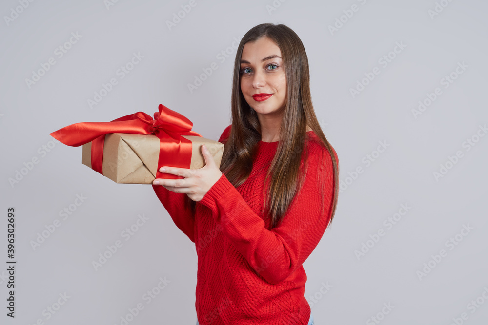 A beautiful young woman in a red sweater, holding a gift box, stands in profile, on a gray background. Holiday concept, Christmas, New Year A beautiful young woman in a red sweater, holding a gift box, stands in profile, on a gray background. Holiday concept, Christmas, New Year