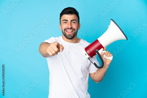 Caucasian man over isolated blue background holding a megaphone and smiling while pointing to the front