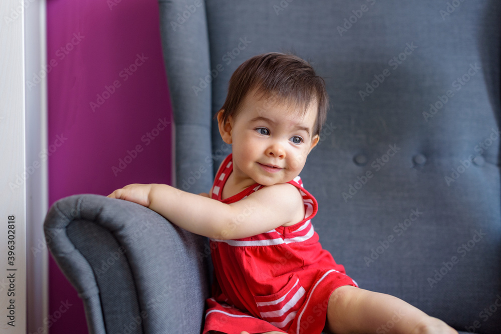 Niña pequeña simpática y sonriente con vestido rojo con lineas blancas sentada en sillón gris Niña pequeña simpática y sonriente con vestido rojo con lineas blancas sentada en sillón gris