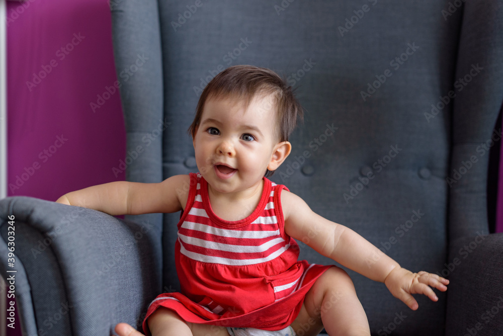 Niña pequeña simpática y sonriente con vestido rojo con lineas blancas sentada en sillón gris Niña pequeña simpática y sonriente con vestido rojo con lineas blancas sentada en sillón gris