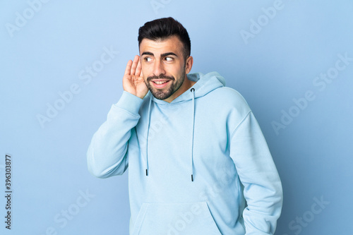 Caucasian man over isolated blue background listening to something by putting hand on the ear