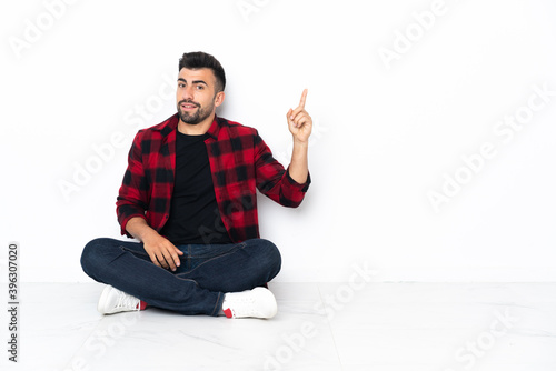 Young handsome man sitting on the floor showing and lifting a finger in sign of the best © luismolinero
