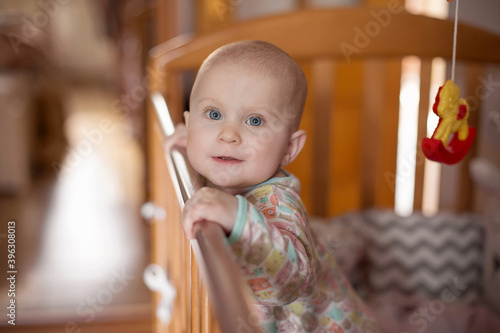 A small six-month-old blue-eyed baby is standing in the crib, holding on to the side. He looks at the camera and smiles. Orange-brown interior in the room. photo