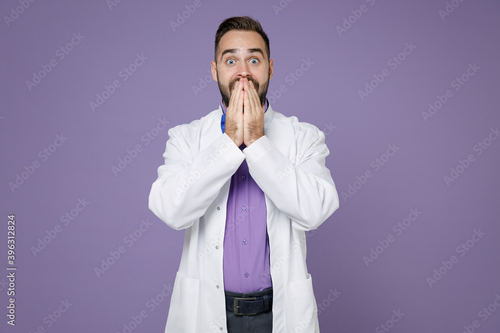 Shocked young bearded doctor man wearing white medical gown covering mouth with hands looking camera isolated on violet colour background studio portrait. Healthcare personnel health medicine concept. Shocked young bearded doctor man wearing white medical gown covering mouth with hands looking camera isolated on violet colour background studio portrait. Healthcare personnel health medicine concept.
