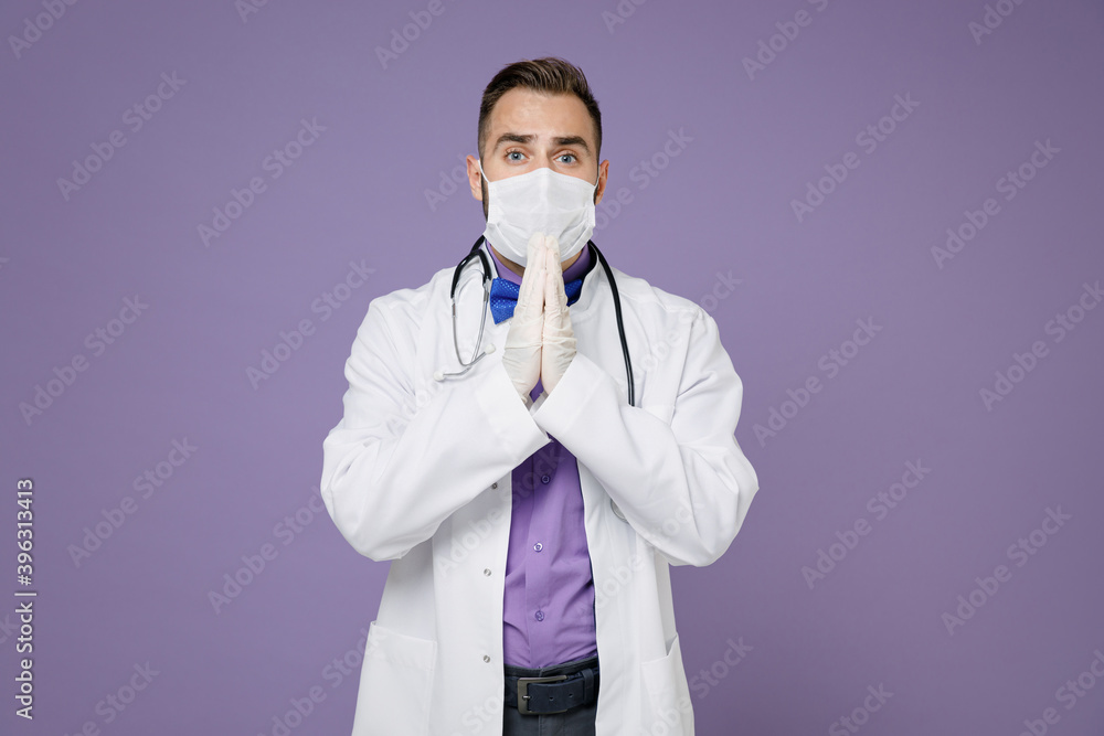Pleading young doctor man in medical gown stethoscope face mask safe from coronavirus virus covid-19 hold hands in prayer isolated on violet background. Healthcare personnel health medicine concept. Pleading young doctor man in medical gown stethoscope face mask safe from coronavirus virus covid-19 hold hands in prayer isolated on violet background. Healthcare personnel health medicine concept.