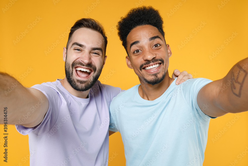 Close up of cheerful young friends european african american men 20s wearing casual violet blue t-shirts doing selfie shot on mobile phone hugging isolated on yellow colour background studio portrait. Close up of cheerful young friends european african american men 20s wearing casual violet blue t-shirts doing selfie shot on mobile phone hugging isolated on yellow colour background studio portrait.
