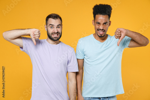 Dissatisfied upset young friends european african american men 20s wearing casual violet blue t-shirts showing thumbs down looking camera isolated on bright yellow colour background studio portrait.