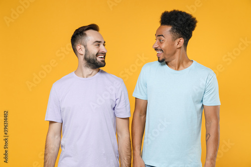 Excited smiling young friends european african american men 20s wearing casual violet blue t-shirts standing looking at each other isolated on bright yellow colour wall background studio portrait.
